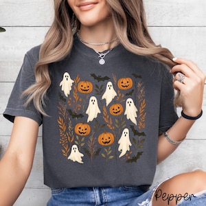 May include: A charcoal grey short-sleeve t-shirt featuring a central graphic print of white ghosts, orange jack-o'-lantern pumpkins, black bats, and brown autumn foliage. The shirt has a crew neckline and a relaxed fit with a vintage-style wash.