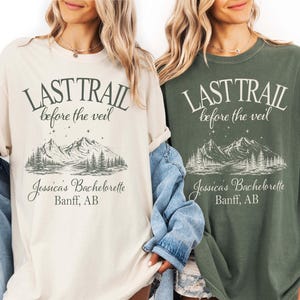 Peut inclure: Deux t-shirts à manches courtes et col rond, l'un crème et l'autre vert foncé, arborant chacun une impression graphique avec le texte "LAST TRAIL before the veil", "Jessica's Bachelorette" et "Banff, AB" au-dessus d'un paysage de montagne.