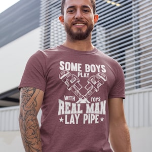 Peut inclure: Un t-shirt à manches courtes bordeaux avec un motif blanc représentant deux clés à pipe croisées. Le texte indique : SOME BOYS PLAY WITH TOYS REAL MAN LAY PIPE. Le design est centré sur la poitrine avec un aspect vieilli et usé.