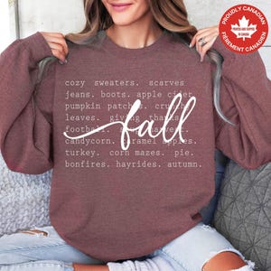 May include: A mauve long-sleeve crewneck sweatshirt featuring white text listing autumn-themed words including cosy jumpers, scarves, jeans, boots, apple cider, pumpkin patches, crunchy leaves, thanksgiving, football, harvest, candy corn, caramel apples, turkey, corn mazes, pie, bonfires, hayrides, and autumn.