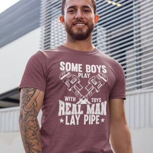 Peut inclure: Un t-shirt à manches courtes bordeaux avec un motif blanc représentant deux clés à pipe croisées. Le texte imprimé sur le devant indique : SOME BOYS PLAY WITH TOYS REAL MAN LAY PIPE, avec deux petites étoiles encadrant la dernière ligne.