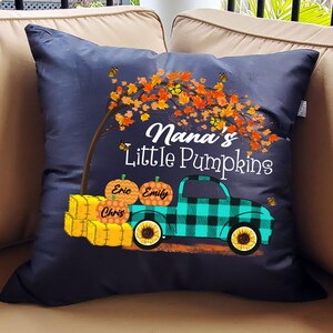 May include: A dark blue throw pillow with a fall-themed design. It features a truck carrying pumpkins labeled "Eric", "Emily", and "Chris". The truck is driving under a tree with orange and yellow leaves. The text "Nana's Little Pumpkins" is printed on the pillow.