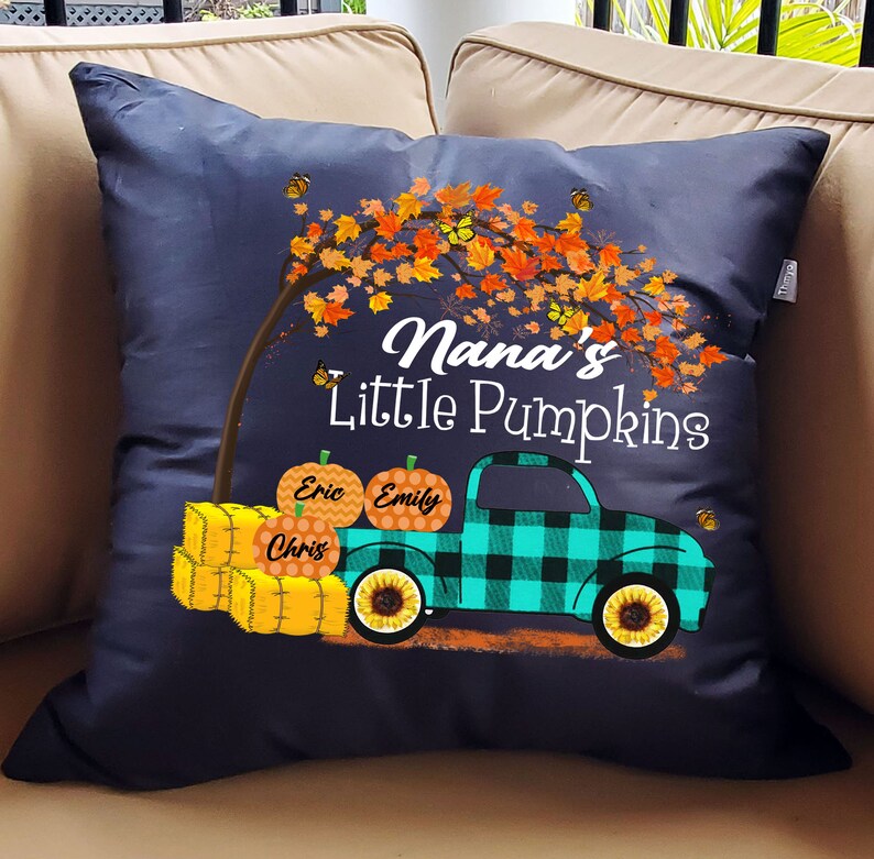May include: A dark blue throw pillow with a fall-themed design. It features a truck carrying pumpkins labeled "Eric", "Emily", and "Chris". The truck is driving under a tree with orange and yellow leaves. The text "Nana's Little Pumpkins" is printed on the pillow.