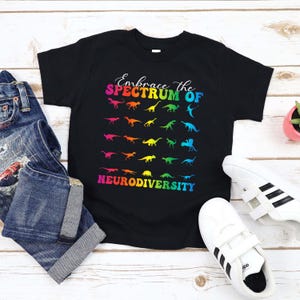 May include: A black short-sleeve t-shirt featuring a multi-colored graphic of various dinosaur silhouettes. The text reads Embrace the SPECTRUM OF NEURODIVERSITY in a rainbow gradient font. The design is centered on the front of the garment.