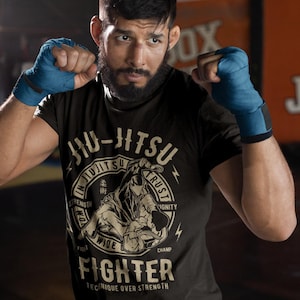 May include: A black short-sleeved t-shirt featuring a beige graphic print of two figures grappling in a circular frame. The text reads JIU-JITSU, IN JIUJITSU WE TRUST, STRENGTH, DIGNITY, FIGHT, WIDE, CHAMP, FIGHTER, and TECHNIQUE OVER STRENGTH.