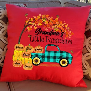 May include: Red throw pillow with a graphic of a teal and black plaid pickup truck carrying pumpkins with names on them. The truck is driving under a tree with orange and yellow leaves. The text "Grandma's Little Pumpkins" is above the truck.