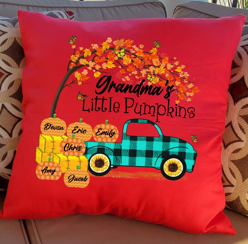 May include: Red throw pillow with a graphic of a teal and black plaid pickup truck carrying pumpkins with names on them. The truck is driving under a tree with orange and yellow leaves. The text "Grandma's Little Pumpkins" is above the truck.