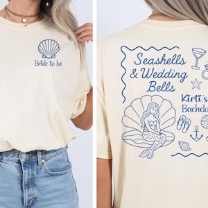 May include: A cream-coloured t-shirt with a blue seashell and the text "Bride to be" on the front. The back displays "Seashells & Wedding Bells" text, a mermaid, and beach-themed illustrations. "Kirti's Bachelorette" is also printed on the back.
