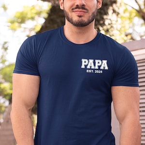May include: A navy blue short-sleeved t-shirt featuring white collegiate-style lettering on the left chest that reads "PAPA" with "EST. 2025" printed directly underneath in a smaller, clean, sans-serif font.