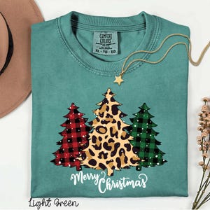 May include: A teal short-sleeve t-shirt with a Comfort Colors label in size XL. The graphic features three stylised Christmas trees: one red buffalo plaid, one leopard print, and one green buffalo plaid, with the text Merry Christmas below.