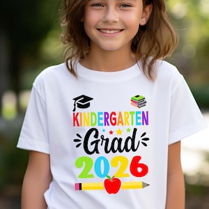 May include: A white t-shirt with a colourful graphic that says "Kindergarten Grad 2026". The graphic includes a graduation cap, a stack of books, stars, and a pencil with an apple.