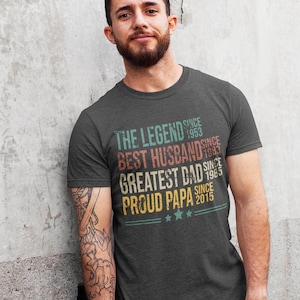 May include: A charcoal gray short-sleeve t-shirt featuring distressed multi-colored text that reads: THE LEGEND SINCE 1953, BEST HUSBAND SINCE 1983, GREATEST DAD SINCE 1985, PROUD PAPA SINCE 2015, with three small stars and horizontal lines at the bottom.
