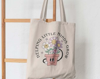 Pode incluir: Uma sacola de lona bege com um gráfico de um vaso de flores rosa com um rosto, contendo três flores sorridentes e um pequeno regador. O texto "HELPING LITTLE MINDS GROW" está impresso em um arranjo circular ao redor da ilustração.