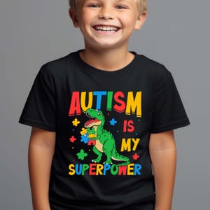 May include: A black t-shirt featuring a colourful graphic of a dinosaur holding a jigsaw piece. The text "AUTISM IS MY SUPERPOWER" is printed on the shirt.