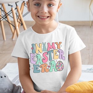 May include: A white short-sleeved t-shirt featuring a multi-coloured graphic print with the text "IN MY EASTER ERA". The design includes a chequered bunny silhouette, a disco ball egg, and small flower accents in pastel shades of pink, blue, yellow, and purple.