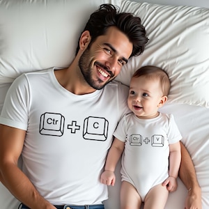 May include: A white short-sleeved t-shirt featuring a black graphic of keyboard keys displaying 'Ctr + C' and a matching white short-sleeved baby bodysuit featuring a black graphic of keyboard keys displaying 'Ctr + V' on the front.