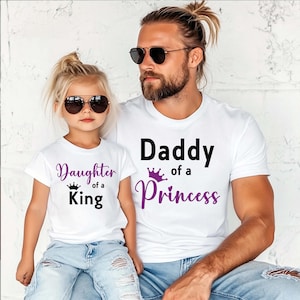 May include: White t-shirts with matching text. One shirt says "Daughter of a King" and the other says "Daddy of a Princess". Both shirts have a crown graphic. The shirts are worn by a father and daughter.