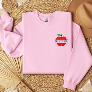 May include: A light pink crewneck sweatshirt featuring a small graphic on the left chest. The graphic depicts a red apple with a green leaf and the text MRS. PETRIELLO printed in black capital letters across the center of the apple design.