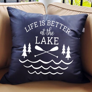 May include: Black throw pillow with white text that reads "Life is better at the lake". The pillow features a graphic of two crossed paddles, two pine trees, and wavy lines representing water.