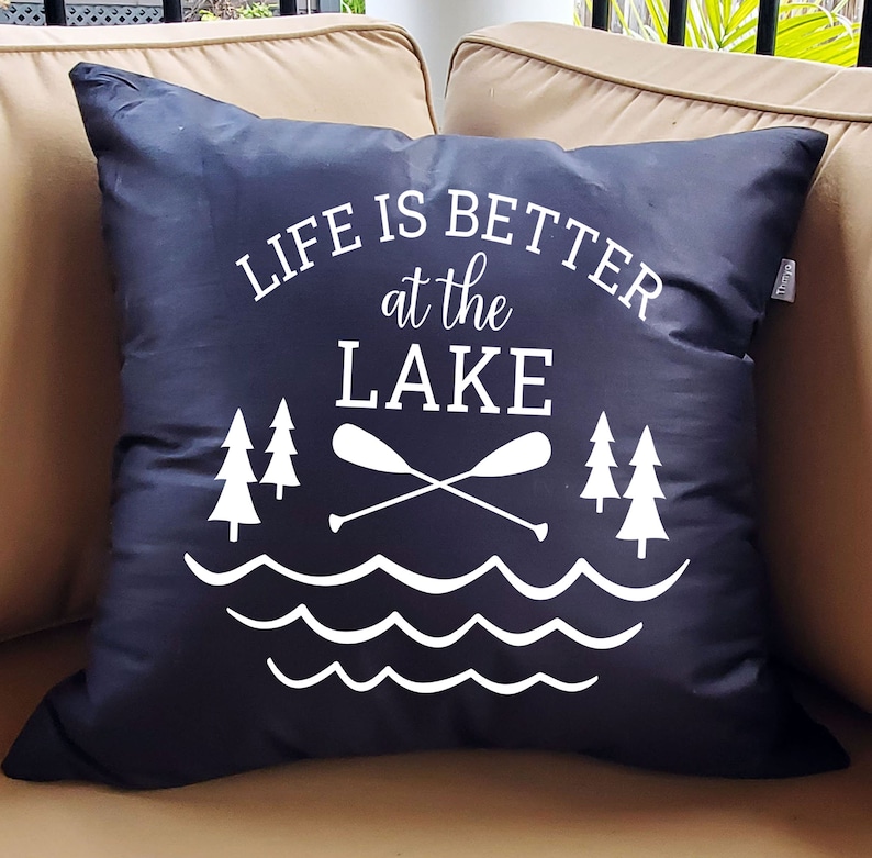 May include: Black throw pillow with white text that reads "Life is better at the lake". The pillow features a graphic of two crossed paddles, two pine trees, and wavy lines representing water.