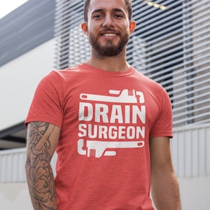May include: A red short-sleeved t-shirt featuring a white graphic print on the chest. The design includes the text DRAIN SURGEON in bold capital letters, flanked by two stylised white pipe wrench illustrations positioned above and below the text.