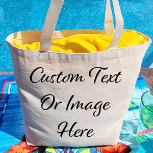 May include: A medium-sized off-white canvas tote bag with two white fabric handles. The front of the bag features the black printed text "Custom Text Or Image Here". A bright yellow towel is tucked inside the bag, which is visible at the top.