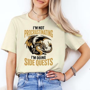 May include: A cream-coloured t-shirt featuring a graphic of a brown and gold dragon in the centre. The text above the dragon reads I'M NOT PROCRASTINATING and the text below the dragon reads I'M DOING SIDE QUESTS in bold, gold-coloured lettering.