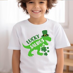 May include: White t-shirt with a green dinosaur wearing a green top hat with a gold buckle. The shirt has the words "LUCKY SAURUS" in green, along with green shamrocks. A fun St. Patrick's Day design.