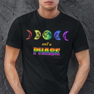 May include: A black short-sleeved t-shirt featuring a graphic of five moon phases in a rainbow gradient. Below the moons, the text reads "not a" in yellow and "PHASE" in a bold, multi-coloured rainbow tie-dye pattern with a black outline.