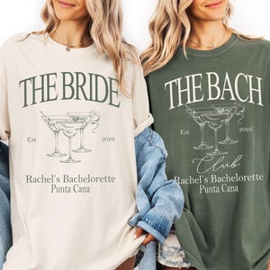 Peut inclure: Deux t-shirts à manches courtes et col rond, un crème et un vert olive, avec du texte vert foncé et des dessins au trait de verres à cocktail. Le t-shirt crème porte l'inscription THE BRIDE, et le vert THE BACH CLUB, avec Est 2026, Rachel's Bachelorette, Punta Cana.