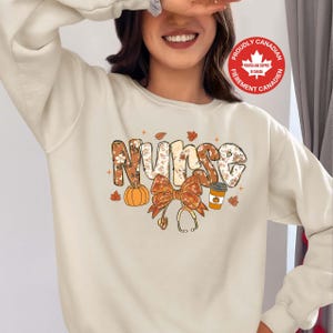May include: Cream-coloured sweatshirt with a fall-themed design. The word "Nurse" is spelled out in a floral and patterned font, accompanied by a pumpkin, bow, coffee cup, and stethoscope. The design includes autumn leaves.