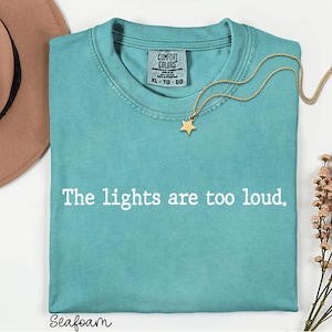 May include: A seafoam green short-sleeve t-shirt with a crew neckline and the white text "The lights are too loud." printed across the chest. The shirt features a Comfort Colors brand label at the inner neck and is size XL.