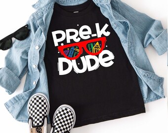 Op de afbeelding: Een zwart t-shirt met de witte tekst PRE-K DUDE en een rode bril met afbeeldingen van schoolspullen in de glazen. Het t-shirt ligt onder een lichtblauw spijkerhemd, vergezeld door een zwarte zonnebril en instappers met een dambordpatroon.