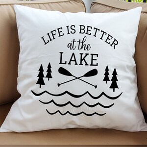 May include: White pillow with black text that reads "Life is better at the lake". The text is surrounded by a graphic of a lake with waves, two crossed paddles, and three pine trees.