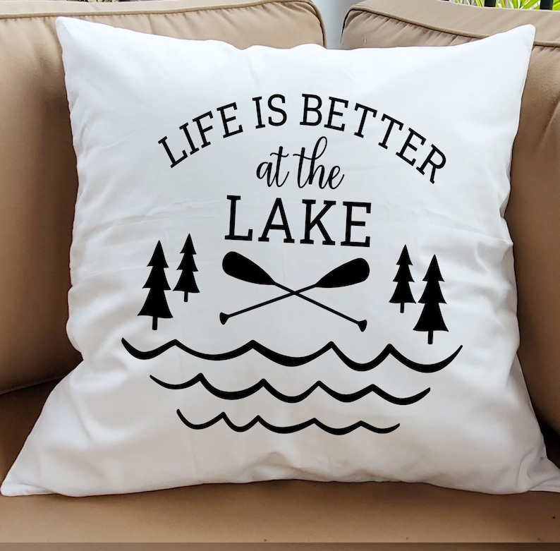 May include: White pillow with black text that reads "Life is better at the lake". The text is surrounded by a graphic of a lake with waves, two crossed paddles, and three pine trees.