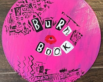 May include: A pink, hand-painted vinyl record with the words "BURN BOOK" in black and white block letters. The record features a red lip design and black scribbled accents.