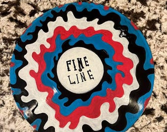 May include: A painted vinyl record with a colorful design. The record features a white center with the words "FINE LINE" in black. The outer design includes red, blue, black, and white wavy patterns. The record is circular and sits on a speckled surface.