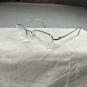 May include: A pair of eyeglasses with a silver metal frame and clear lenses. The glasses have a rectangular shape and are resting on a white surface. The temples are thin and extend back to the ears.