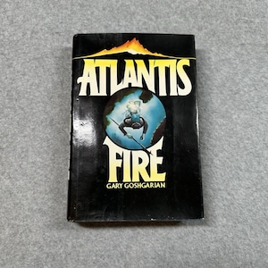 Op de afbeelding: Een hardcover boek met de titel "Atlantis Fire" van Gary Goshgarian. De omslag is zwart met de titel in wit en geel, en een illustratie van een duiker. Het boek is in goede staat.