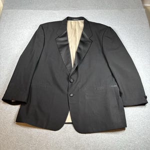 Op de afbeelding: Zwart smokingjasje met satijnen revers en een sluiting met twee knopen. De jas heeft twee voorzakken en een beige voering. De mouwen hebben knopen bij de manchetten. Het kledingstuk ligt plat op een grijze ondergrond.