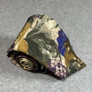 May include: Rolled necktie with a floral pattern in shades of blue, brown, green, and purple. The tie is made of a silky material and is rolled up, showcasing the pattern.
