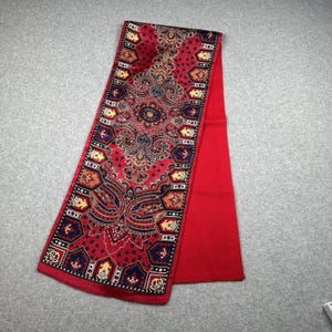 May include: A red scarf with a detailed paisley pattern in shades of gold, navy, and burgundy. The scarf has a solid red border and a rectangular shape. The intricate design is centered on the scarf.