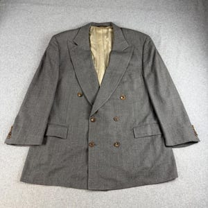 May include: A double-breasted blazer in a medium gray fabric with a subtle vertical pinstripe pattern. The garment features wide lapels, six brown buttons on the front, two flap pockets, and a smooth, light tan interior lining visible at the neck.