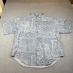 Pode incluir: Camisa azul clara de manga curta com um padrão repetido de formas geométricas e desenhos florais em branco e azul escuro. A camisa tem uma gola clássica e botões de madeira.