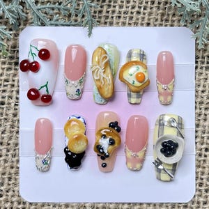 May include: A set of ten artificial nails with various food-themed designs. The nails feature cherries, bread, eggs, and caviar, with pink and patterned backgrounds. Some nails have decorative accents.