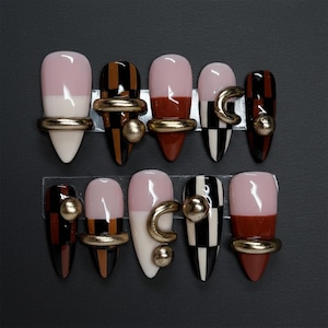 May include: A set of ten artificial nails in various designs. Some nails are half pink and white, others feature brown and black stripes, and some have gold ring and ball accents. The nails are displayed on a clear plastic holder against a black background.