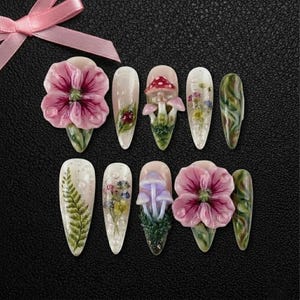 May include: A set of ten artificial nails with various designs. Some nails feature 3D floral and mushroom embellishments, while others showcase fern and floral patterns. The color palette includes pink, green, white, and purple, set against a black background.