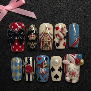 May include: A collection of ten artificial nails with various designs. The nails feature a red polka dot bow, a circus tent, playing card motifs, and a bunny holding balloons. Other designs include a crown, a rose, and a jester.