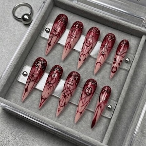 May include: A set of ten stiletto-shaped press-on nails featuring a gradient design that transitions from a nude base to a deep burgundy tip. The nails are adorned with intricate black line art patterns and a dark red glitter finish at the tips.