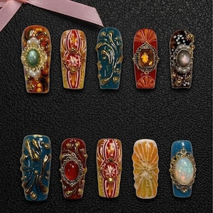 May include: A collection of ten ornate, coffin-shaped artificial nails. Each nail features a unique design with jewel-like embellishments, gold accents, and intricate patterns in shades of red, blue, orange, and brown. The nails are arranged on a dark surface.
